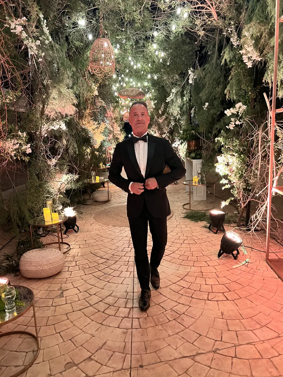 A man in a tuxedo on a garden path under string lights